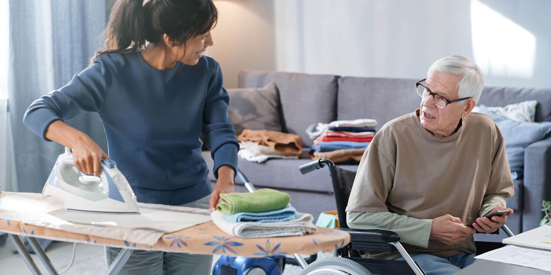 Eine Haushaltshilfe bei Pflegegrad 2 unterstützt einen älteren Mann im Rollstuhl zuhause. Die Pflegekraft bügelt Wäsche und kümmert sich um den Haushalt, während sie freundlich mit dem Senior kommuniziert. Im Hintergrund sind gefaltete Wäschestücke und ein Staubsauger zu sehen – typische Aufgaben der häuslichen Pflege. Diese Aufnahme veranschaulicht das Angebot der Caritas Rhein-Kreis Neuss: professionelle Haushaltshilfe für Pflegebedürftige mit Pflegegrad 2, die ein selbstständiges Leben zuhause ermöglicht.