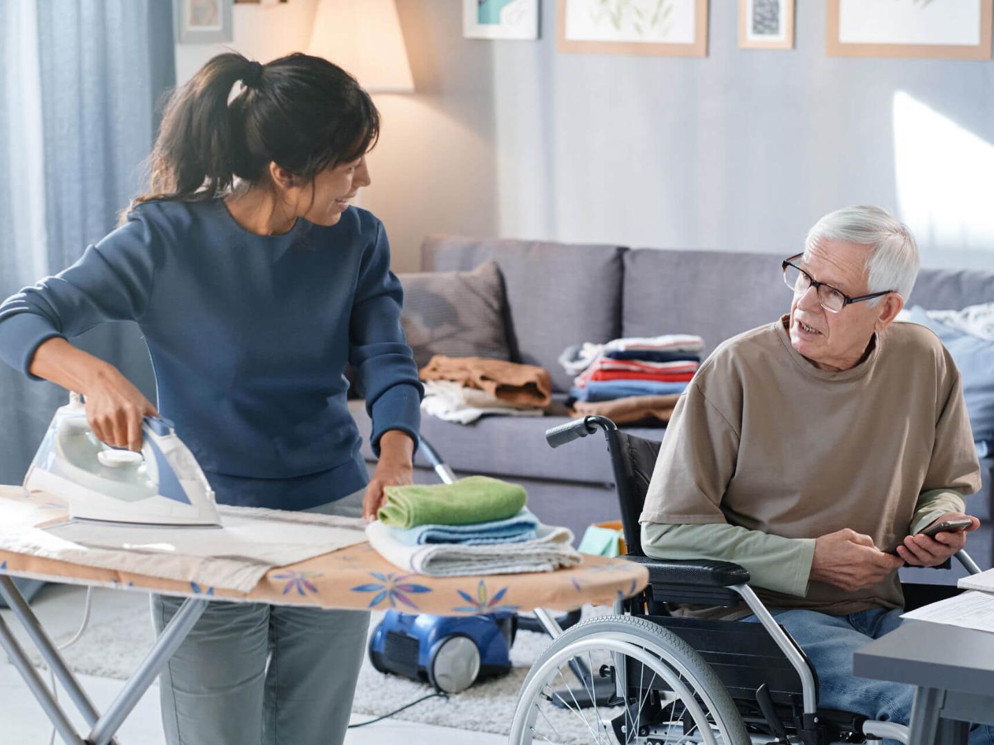 Eine Haushaltshilfe bei Pflegegrad 2 unterstützt einen älteren Mann im Rollstuhl zuhause. Die Pflegekraft bügelt Wäsche und kümmert sich um den Haushalt, während sie freundlich mit dem Senior kommuniziert. Im Hintergrund sind gefaltete Wäschestücke und ein Staubsauger zu sehen – typische Aufgaben der häuslichen Pflege. Diese Aufnahme veranschaulicht das Angebot der Caritas Rhein-Kreis Neuss: professionelle Haushaltshilfe für Pflegebedürftige mit Pflegegrad 2, die ein selbstständiges Leben zuhause ermöglicht.