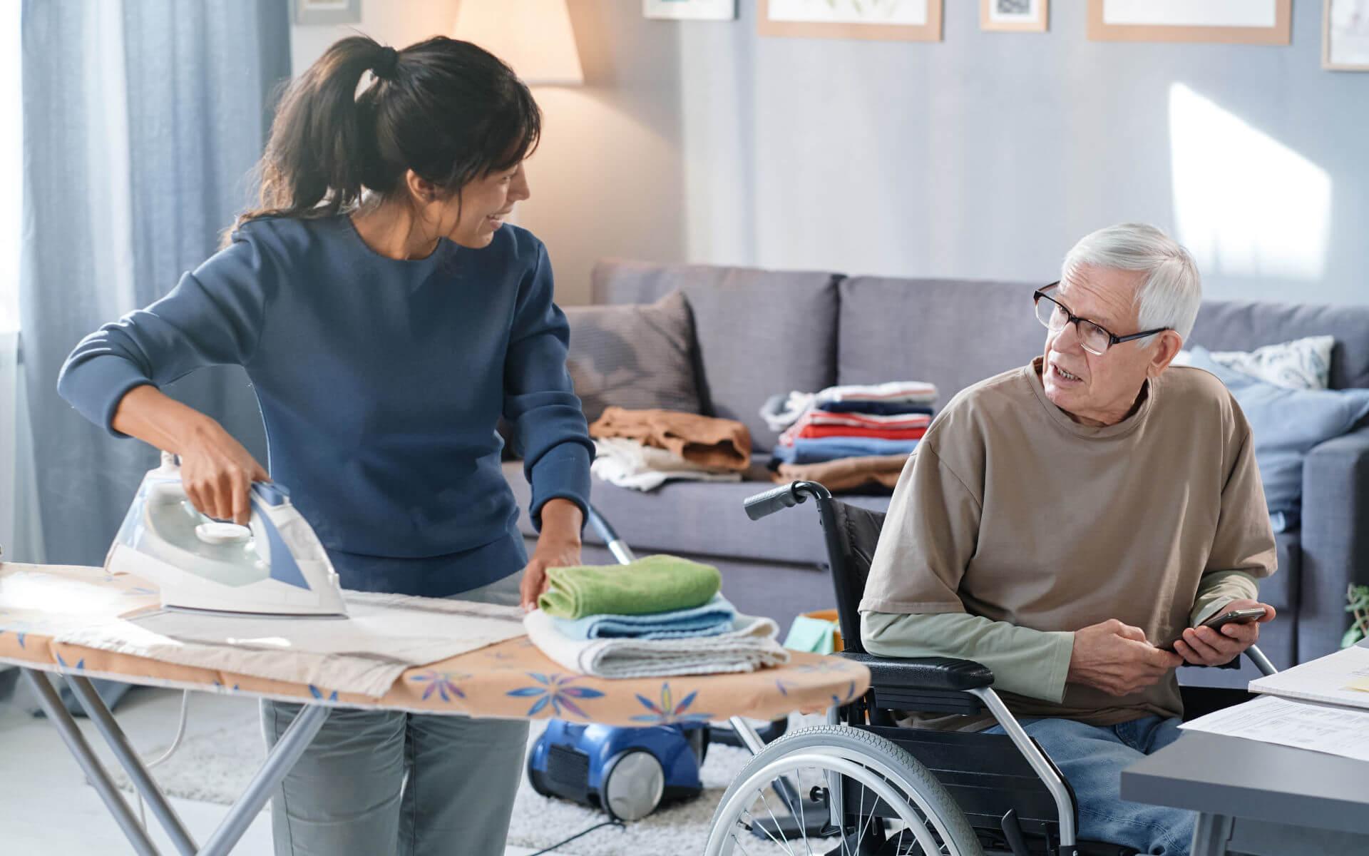 Eine Haushaltshilfe bei Pflegegrad 2 unterstützt einen älteren Mann im Rollstuhl zuhause. Die Pflegekraft bügelt Wäsche und kümmert sich um den Haushalt, während sie freundlich mit dem Senior kommuniziert. Im Hintergrund sind gefaltete Wäschestücke und ein Staubsauger zu sehen – typische Aufgaben der häuslichen Pflege. Diese Aufnahme veranschaulicht das Angebot der Caritas Rhein-Kreis Neuss: professionelle Haushaltshilfe für Pflegebedürftige mit Pflegegrad 2, die ein selbstständiges Leben zuhause ermöglicht.
