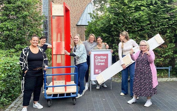 Das Team der Erziehungs- und Familienberatungsstelle Dormagen öffnet auf dem Platz vor der Pfarrkirche  St. Michael beim Netzwerktreffen die rote Tür. Knieend von links: Maren Roskam, Ida Dietrich; hinter der Tür von links: Ines Standura, Daniela Logtenberg, Daniela Schöpe, Sonja Malandrino; rechts neben der Tür:  Claudia Breuer.