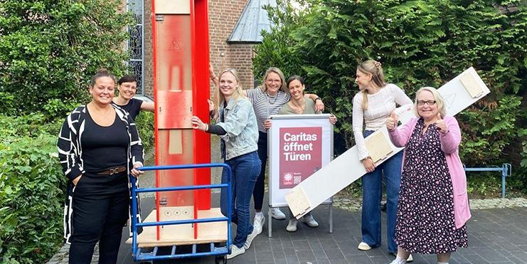 Das Team der Erziehungs- und Familienberatungsstelle Dormagen öffnet auf dem Platz vor der Pfarrkirche St. Michael beim Netzwerktreffen die rote Tür. Knieend von links: Maren Roskam, Ida Dietrich; hinter der Tür von links: Ines Standura, Daniela Logtenberg, Daniela Schöpe, Sonja Malandrino; rechts neben der Tür: Claudia Breuer.