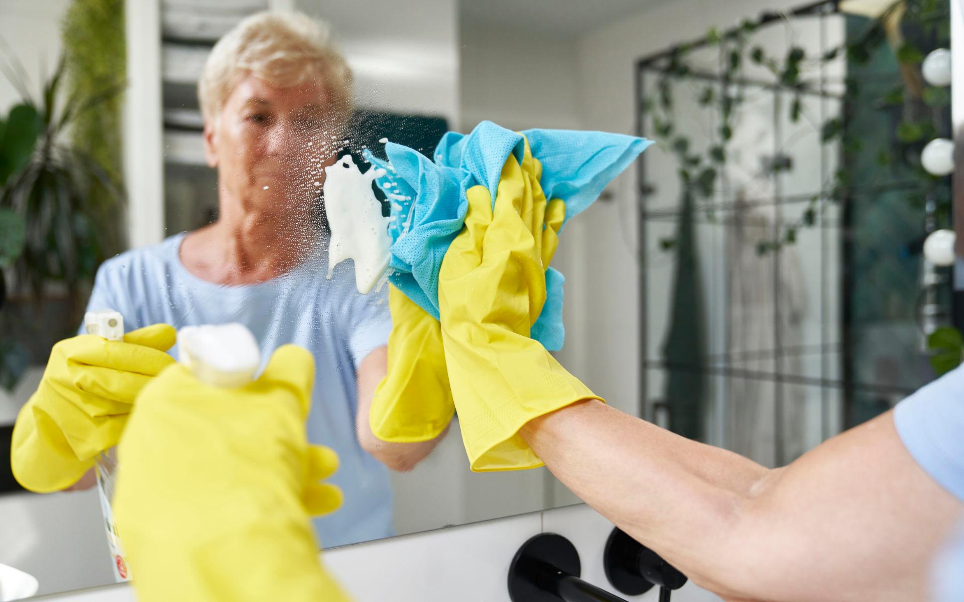 Haushaltshilfe putzt Spiegel bei einem Menschen, der über seinen Pflegegrad 1, die Haushaltshilfe über die Pflegekasse in Anspruch nimmt.